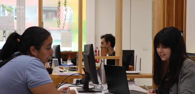  The image shows an individual consultation at the Valencia Energy Office. On the left, a woman is sitting at a desk speaking with another woman on the right, who is smiling while holding a pen and reviewing a document. Both are seated in front of computers in a modern, open-space office setting with light wooden elements and plants hanging from above, creating a welcoming environment. The consultation is focused on energy-related advice.