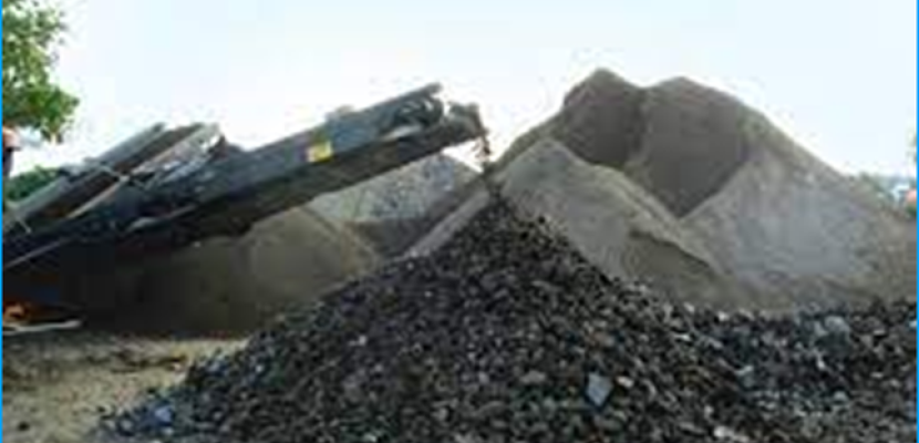 A mechanical crusher discharges a pile of mixed aggregates, featuring both larger dark, rocky materials in the foreground and finer sandy aggregates in the background. The machine's conveyor is visible to the left, depositing crushed materials, with large heaps of sand and gravel behind it. This scene depicts the process of crushing and sorting raw materials into different grades for use in construction. Trees and natural surroundings frame the industrial activity.