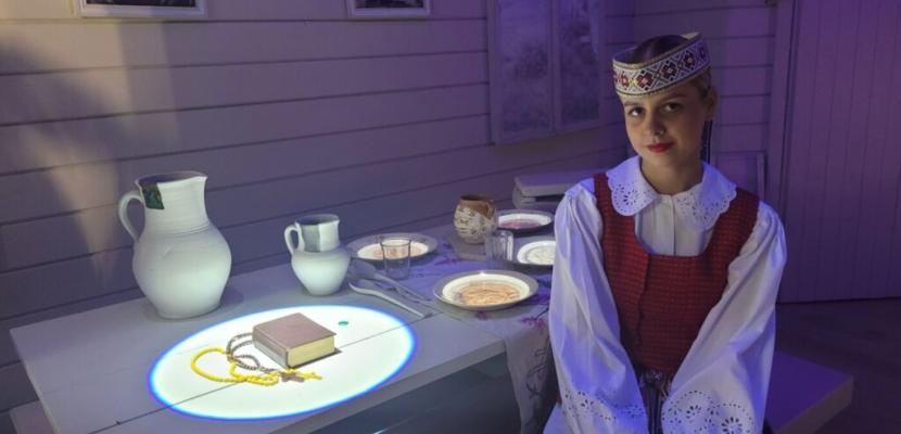 This image represents an interactive cultural display at the Butrimonys museum, illustrating the everyday life and religious practices of local communities through digital projection. The image shows a woman dressed in traditional Lithuanian attire, seated beside a table with a projection of religious items such as a book and rosary beads. The table is part of an interactive exhibit, with projected images of traditional dishes, objects. In the background, religious icons and a cross are visible on the wall.