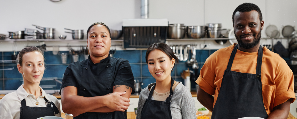 Picture of 4 people from migrant background, 3 women and a men in a kitchen, learning the skills to work in a kitchen, smiling