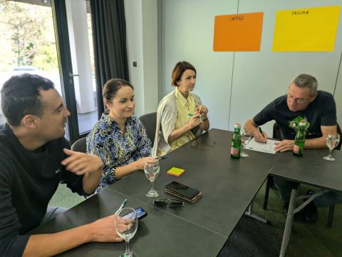 A group of four individuals participating in a discussion at a meeting. They are seated at a table with water bottles, glasses, and documents in front of them. Two posters labeled "Snage" (Strengths) and "Prilike" (Opportunities) are visible on the wall in the background.