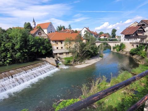 A scenic view of a waterfall flowing into a river in Škofja Loka, Slovenia, with historic buildings and a stone bridge in the background. The red-tiled roofs and lush greenery complement the serene blue sky, creating a picturesque landscape.