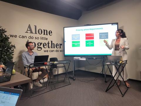 A woman stands presenting in front of a large screen displaying priority areas and departments, while a man sits nearby with a laptop. The room features a motivational quote on the wall that reads, "Alone we can do so little; together we can do so much.