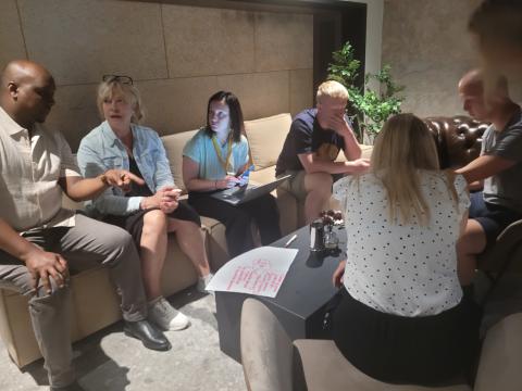 A diverse group of people sitting on sofas and chairs in a cozy room, actively engaged in a discussion about stakeholder engagement. One person is using a laptop, while another is taking notes on a piece of paper with brainstorming ideas.