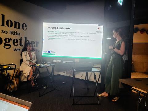 Two women are presenting in a conference room. One is standing and speaking in front of a screen displaying "Expected Outcomes," while the other is seated, listening attentively. The wall features a motivational quote that reads, "Alone we can do so little; together we can do so much."