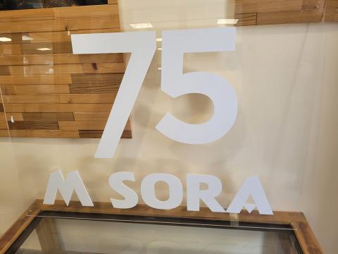 A display sign reading "75 M SORA" against a wooden background, showcasing the company's 75th anniversary.