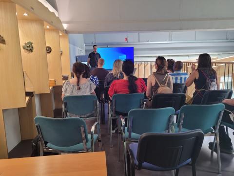 A group of people seated in a room with wooden displays, listening to a presentation by a speaker standing in front of a large screen.