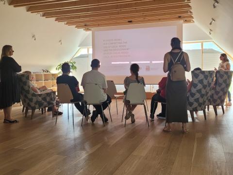 A group of people seated in a well-lit room with wooden floors and ceiling beams, watching a presentation on a large screen about the business model for connecting the use of local wood and developing high-value-added wood products.