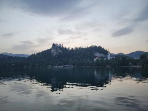 A serene view of Lake Bled at dusk, with Bled Castle perched on a hill and a church visible near the shoreline, reflecting in the calm water.