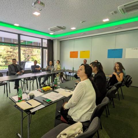 A group of individuals seated around a U-shaped table during a meeting, attentively listening to a presentation. Sticky note posters labeled "Snage" (Strengths), "Prilike" (Opportunities), "Slabosti" (Weaknesses), and "Prijetnje" (Threats) are visible on the wall, representing a SWOT analysis discussion.