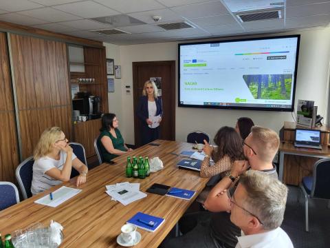 Group of people seated around a conference table, listening to a woman presenting. A large screen in the background displays the presentation.