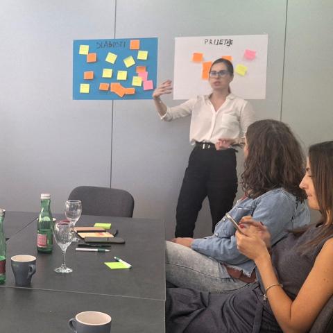 A woman stands in front of a board with colorful sticky notes during a meeting, explaining points related to weaknesses and threats in a SWOT analysis. Two other participants are seated at the table, listening attentively. Posters on the wall are labeled "Slabosti" (Weaknesses) and "Prijetnje" (Threats).