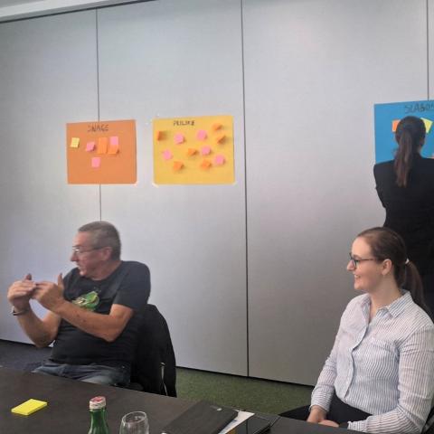 Two participants engage in discussion during a meeting, with colorful sticky note boards in the background labeled "Snage" (Strengths), "Prilike" (Opportunities), and "Slabosti" (Weaknesses) as part of a SWOT analysis. One participant is writing on the board while others listen.