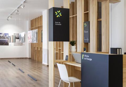 The image shows a clean and modern interior of the Energy Office located in the Ayora neighbourhood of Valencia. The space features light wooden structures and minimalist design, with the "Oficina de l'Energia" signage visible on a dark counter in the foreground. Above the counter, there's a sign that reads "Punt de Connexió" (Connection Point). The wooden accents and bright lighting create a welcoming atmosphere for visitors seeking energy-related advice and resources.