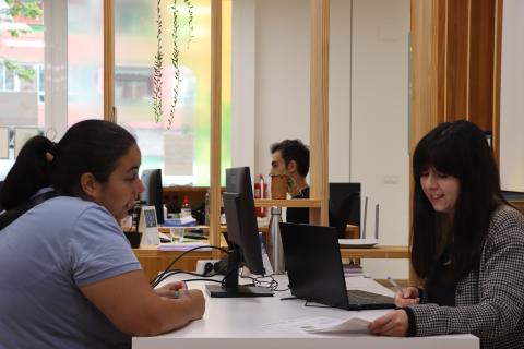  The image shows an individual consultation at the Valencia Energy Office. On the left, a woman is sitting at a desk speaking with another woman on the right, who is smiling while holding a pen and reviewing a document. Both are seated in front of computers in a modern, open-space office setting with light wooden elements and plants hanging from above, creating a welcoming environment. The consultation is focused on energy-related advice.