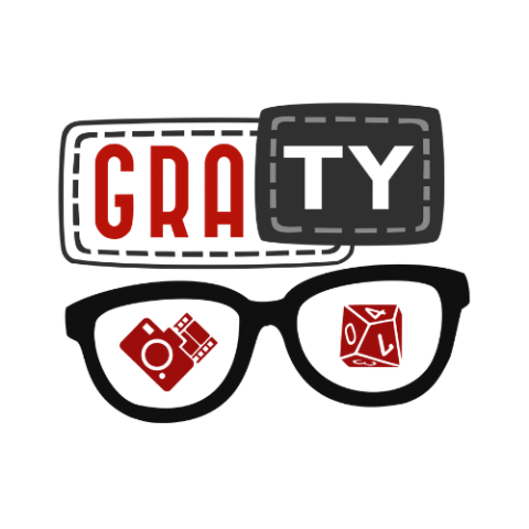 The image shows the logo of the GraTY Foundation. It consists of two rectangular boxes: one with the letters "GRA" in red and the other with "TY" in white on a black background. Below these boxes is a stylized pair of glasses, with one lens containing a red circuit board and the other featuring a red die. This logo reflects the foundation's focus on games, technology, and creative activities.