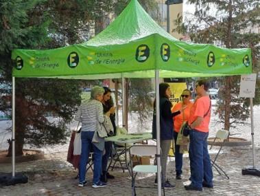 The image shows the mobile Energy Office of Valencia, set up outdoors under a bright green tent with the "Oficina de l'Energia" logo displayed on the canopy. A small group of people is gathered around the table inside the tent, receiving energy-related advice and information from staff members. The setup provides a temporary, accessible point for citizens to engage with energy experts in various parts of the city, promoting sustainability and energy efficiency through direct community interaction. 