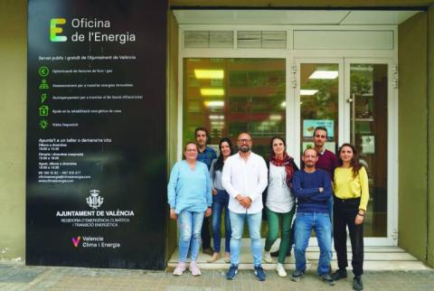 Part of the Energy Office Team, in front of the Ayora Energy Office in Valencia. The team is diverse and welcoming. A sign next to the Office door shows the main characteristics of this public, free service of Valencia City Council. 