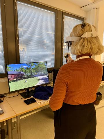 A person wearing VR headgear, holding a virtual hand controller, standing in front of a computer monitor.