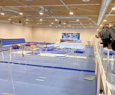 A large gymnastics hall with a blue floor and various gymnastics apparatus, and a spectators gallery.