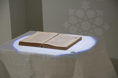 The image shows an open Quran displayed on a white cloth-covered table. The Arabic text on the pages is visible, and a spotlight illuminates the holy book. Next to the book is a button, which serves an interactive purpose, allowing visitors to listen to Quranic recitations or learn more about Islamic traditions. 