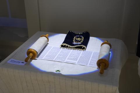 The image shows a Jewish Torah scroll displayed on a table, accompanied by a decorative blue and gold velvet Torah cover. The scroll is partially unrolled, revealing a portion of Hebrew text. On the right side, pressing the button allows visitors to hear traditional Jewish prayers.