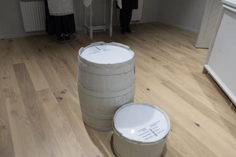 The image shows two white barrels of varying sizes on a wooden floor. The tops of the barrels contain interactive buttons and text as part of a quiz game, where visitors are prompted to answer questions. This interactive game engages visitors by testing their knowledge about local traditions, food, or cultural practices.