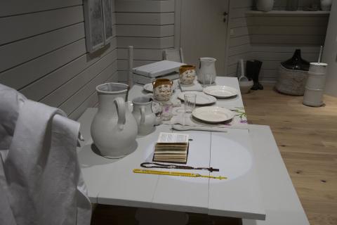 The image shows a traditional dining table set with ceramic jugs, plates, glasses, and utensils, evoking a typical rural Lithuanian household. A book and rosary beads suggest a religious context. By pressing the button, visitors can listen to Christian prayers in Lithuanian, and when the light goes out, traditional Lithuanian dishes are projected onto the white plates, enhancing the interactive experience.