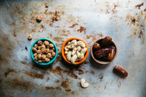 Three bowls on a rustic surface, filled with tiger nuts, cashew nuts, and dates.