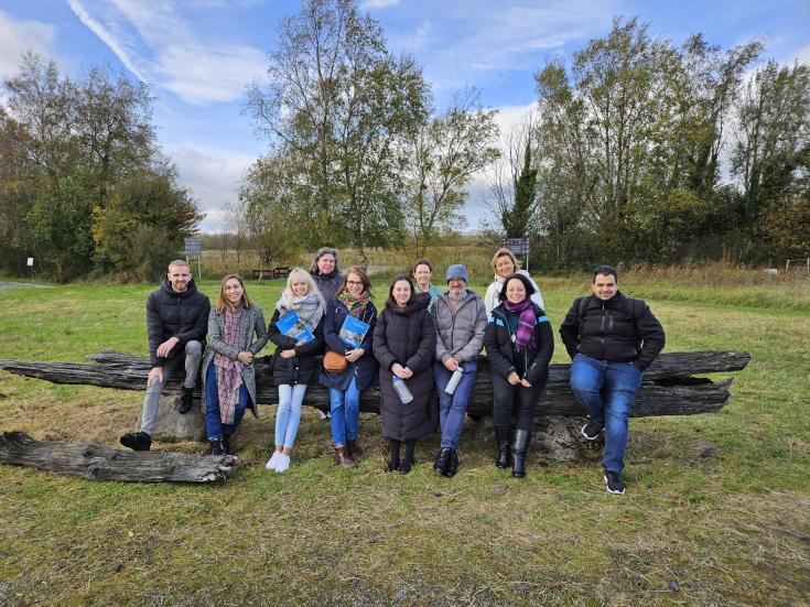 A group of ADAPTO project partners sits together on a large wooden bench outdoors at Cabragh Wetlands, Ireland. Behind them are green trees and an open field. The group, dressed warmly for autumn, is smiling and holding informational brochures.