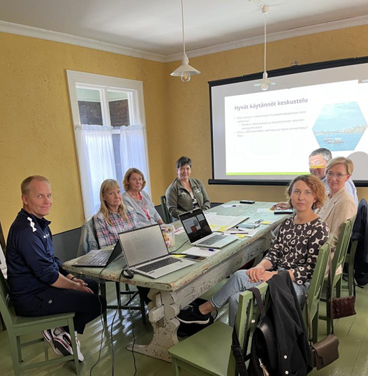 Participants of DETOCS Regional Stakeholder Meeting sit at a table with laptops and a presentation