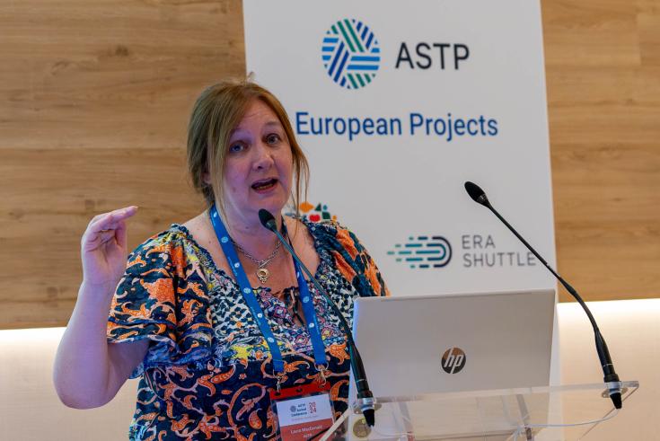 Portrait of ASTP CEO Laura McDonald Speaking at ASTP Annual Conference. In the background there is a banner with all the EU projects ASTP is part of. Laura has a laptop with a michrophone in front of her, and she is looking directly in the camera.