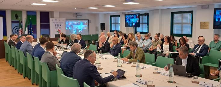 People seated at long meeting table. More people sit nearby and a videoscreen shows additional attendees connecting remotely