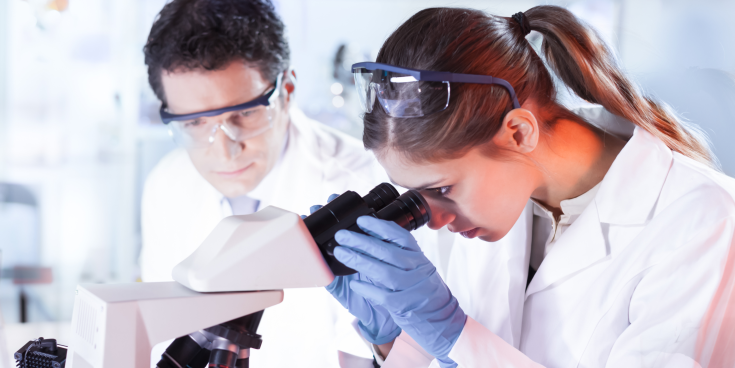 Two scientists ( a man on the left and a woman on the right) in white coats looking in a microscope.