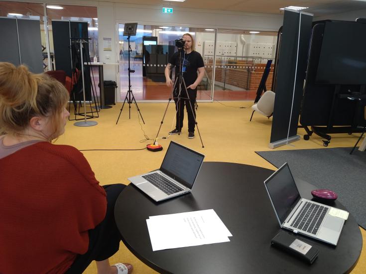 In the background, the cameraman of the video records a woman sitting in front, who speaks while looking at the text on the computer screen.