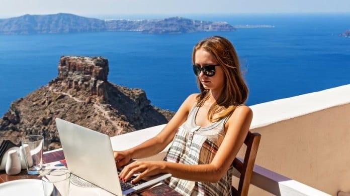 A young woman working on a laptop with sunglasses and vest top on, sitting at a table on a balcony in the sun, overlooking a deep blue body of water with mountain land in the distance.