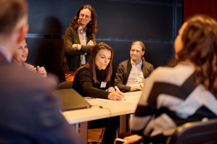 Person writing on paper with people standing around