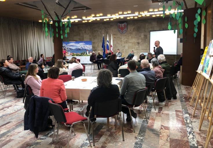 Participants of Kranj's "Ideas Café for a Green Future" gather in the City Hall's atrium, sitting at round tables and engaging in discussions. The event, part of the fourth Kranj Eco Week and the Interreg Europe GIFT project, features a speaker addressing the audience while presentations and posters are displayed. The room is decorated with green leaf cutouts hanging from tree-shaped structures, creating a vibrant and collaborative atmosphere.