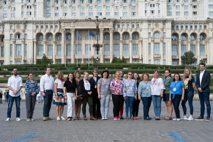 A group of 20 people, composed of both men and women, is standing outside in front of a large, historic building with ornate architecture. They are posing for a group photo in a cobblestone square. Most of the participants are wearing casual or business-casual attire, and several have name badges around their necks, suggesting they are part of an official event or conference.