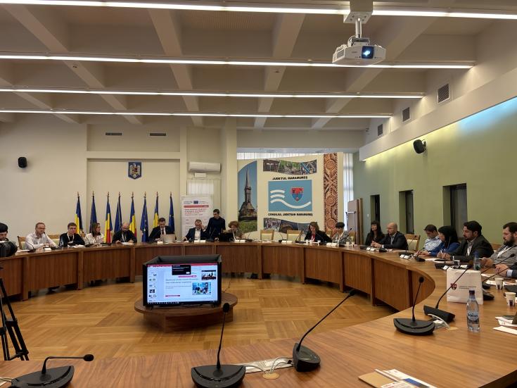 People sitting in a large room at desks arranged in a circle, with a presentation projected on a display in the centre.