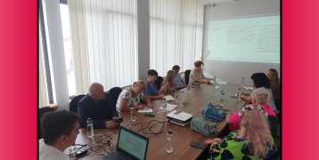 Meeting participants seated around a table in a conference room during a presentation, projected in the background.