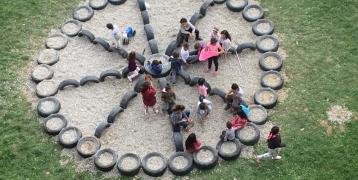 The image shows an outdoor play area where a group of children are gathered. The play area is designed in a circular pattern using old tires, which are arranged in a structured way—possibly resembling a flower or a peace sign. The inside of the structure is filled with gravel or small stones. The children are actively engaging with the space, some sitting on the tires while others stand or walk around. The setting appears to be a grassy field, and the atmosphere is lively and interactive.