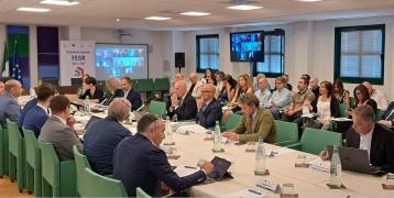 People seated at long meeting table. More people sit nearby and a videoscreen shows additional attendees connecting remotely