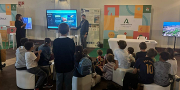 A man lectures in front of a large screen to a group of schoolchildren sitting on chairs and stools.