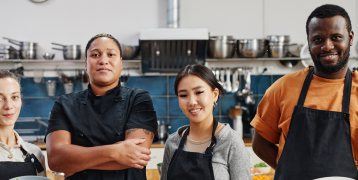 Picture of 4 people from migrant background, 3 women and a men in a kitchen, learning the skills to work in a kitchen, smiling