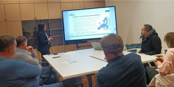 People in a meeting sitting around a table listening to a woman giving a presentation