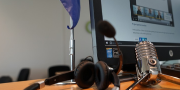 Close-up of a desk setup for virtual meetings or online collaboration. The desk features a European Union flag, a computer monitor displaying a video call or webinar interface, a pair of black headphones, and a vintage-style microphone.