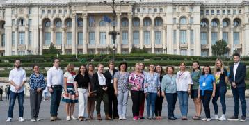 A group of 20 people, composed of both men and women, is standing outside in front of a large, historic building with ornate architecture. They are posing for a group photo in a cobblestone square. Most of the participants are wearing casual or business-casual attire, and several have name badges around their necks, suggesting they are part of an official event or conference.