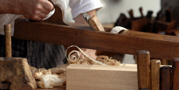 A carpenter's hands using a wooden hand plane to smooth a piece of wood on a workbench, surrounded by wood shavings and various woodworking tools.