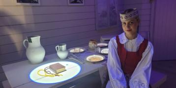 This image represents an interactive cultural display at the Butrimonys museum, illustrating the everyday life and religious practices of local communities through digital projection. The image shows a woman dressed in traditional Lithuanian attire, seated beside a table with a projection of religious items such as a book and rosary beads. The table is part of an interactive exhibit, with projected images of traditional dishes, objects. In the background, religious icons and a cross are visible on the wall.